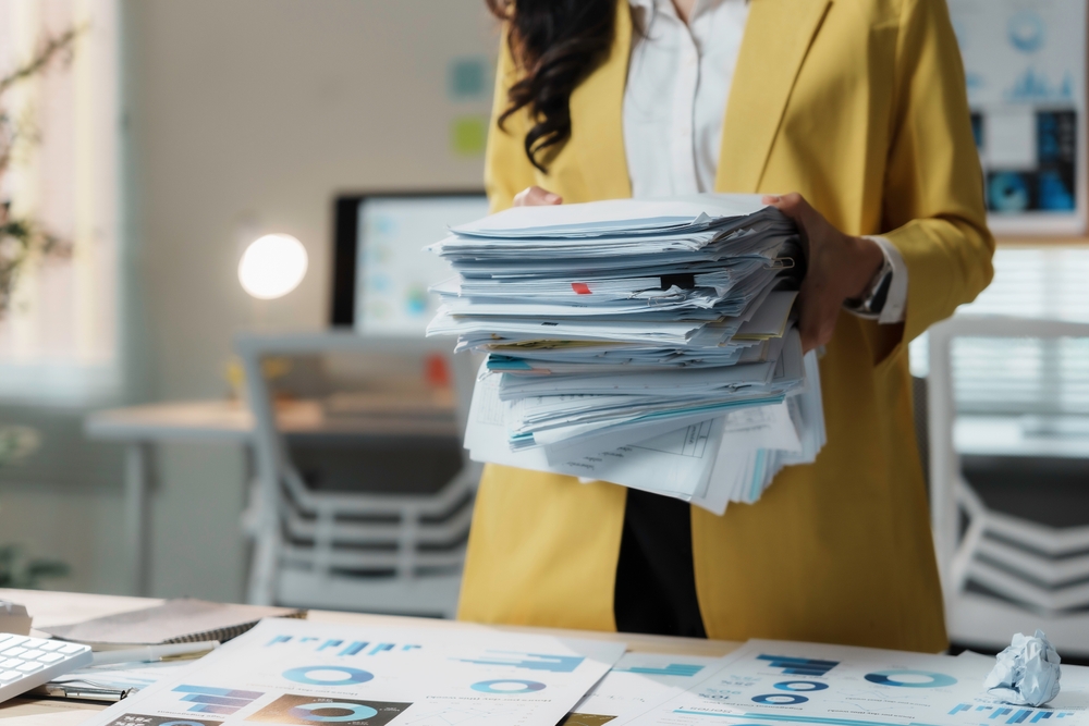 Person carrying a large stack of disorganized paper records and reports across an office