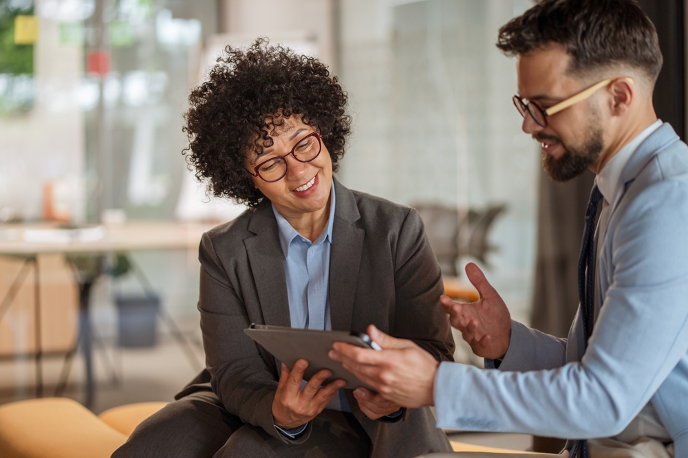 Two professionals in conversation, one holding a tablet while the other explains a point with an open hand.