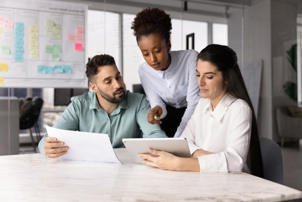 Three colleagues comparing information on a printed document and a tablet screen side by side during a working session.