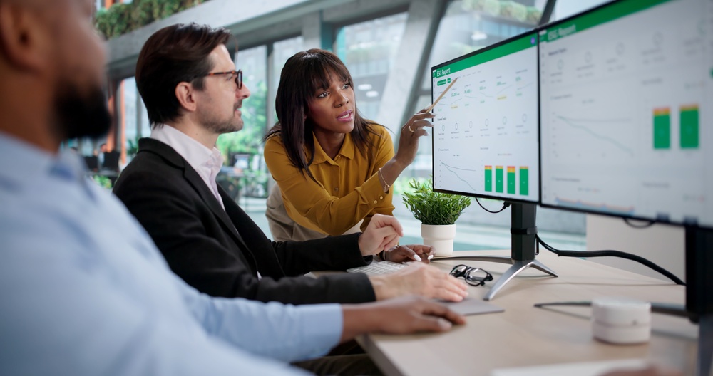 Team reviewing analytics dashboards on dual monitors, one person pointing to a chart while discussing channel performance data.