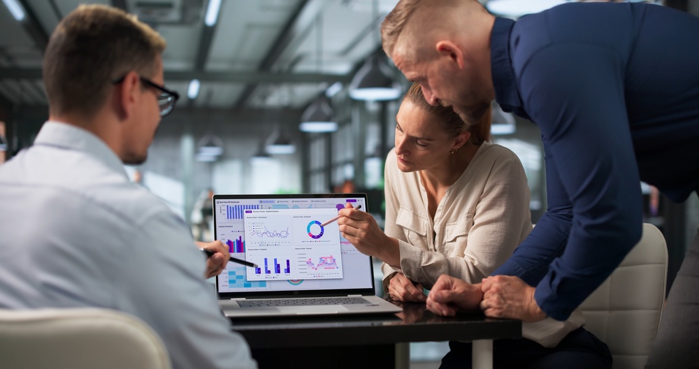Three colleagues reviewing a marketing analytics dashboard on a laptop, discussing campaign performance figures.