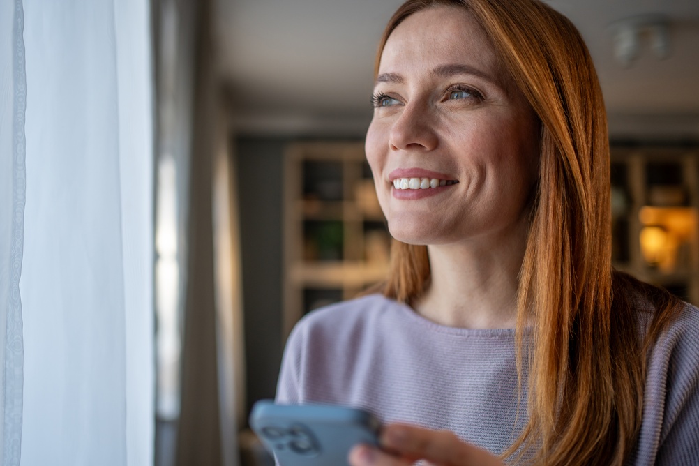 Woman standing at a sunlit window holding a phone and smiling after a call with a behavioral health practice