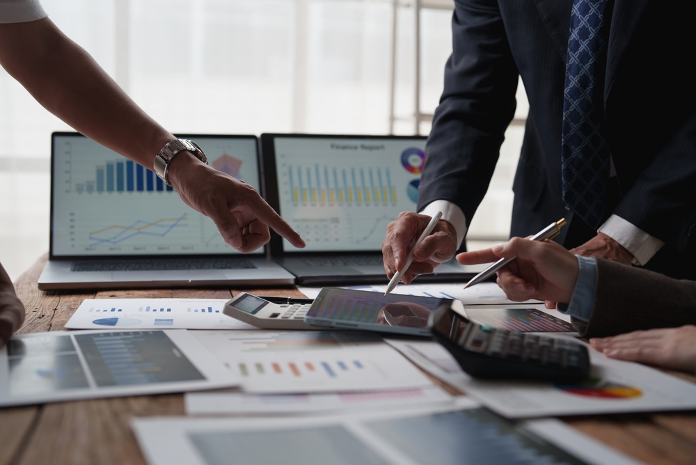 Hands pointing at printed financial reports and charts on a desk with calculators, a tablet, and two laptops showing data dashboards.