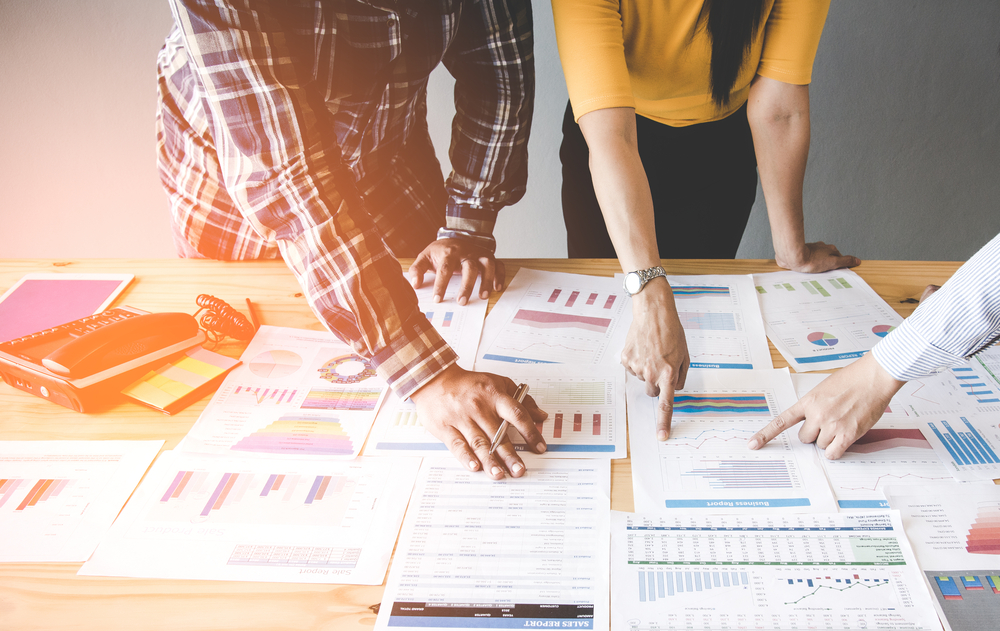 Two colleagues leaning over a table covered in printed sales and business reports, pointing at bar charts as they compare figures.