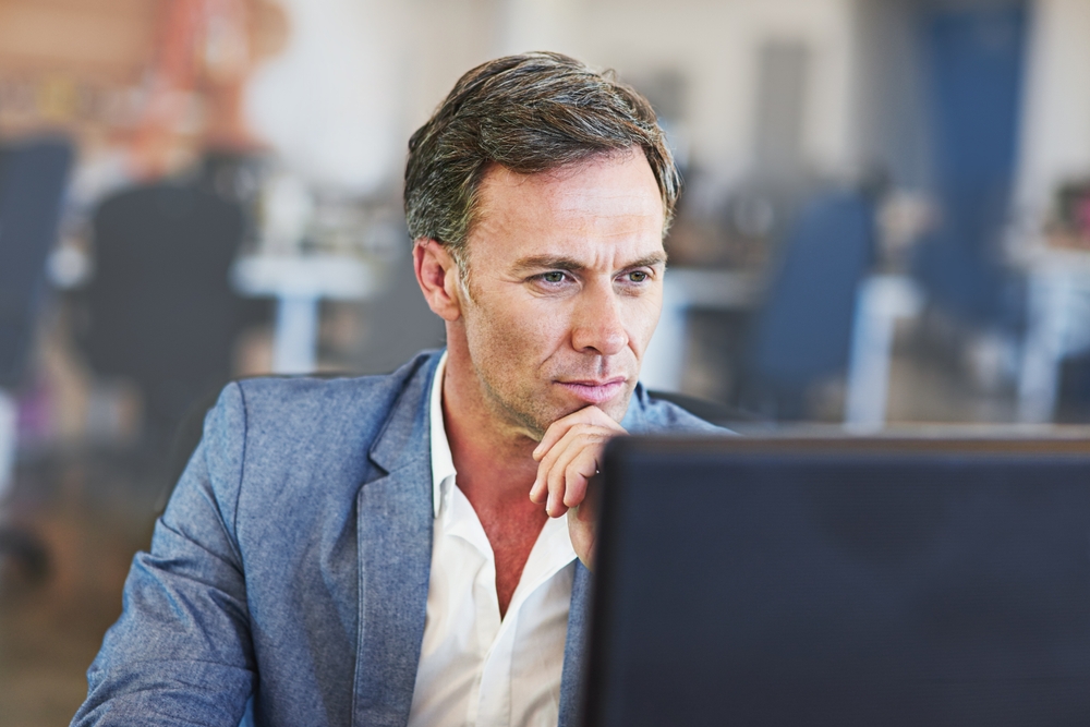 Man in a gray blazer studying a computer screen in an office, hand on his chin, looking thoughtful.