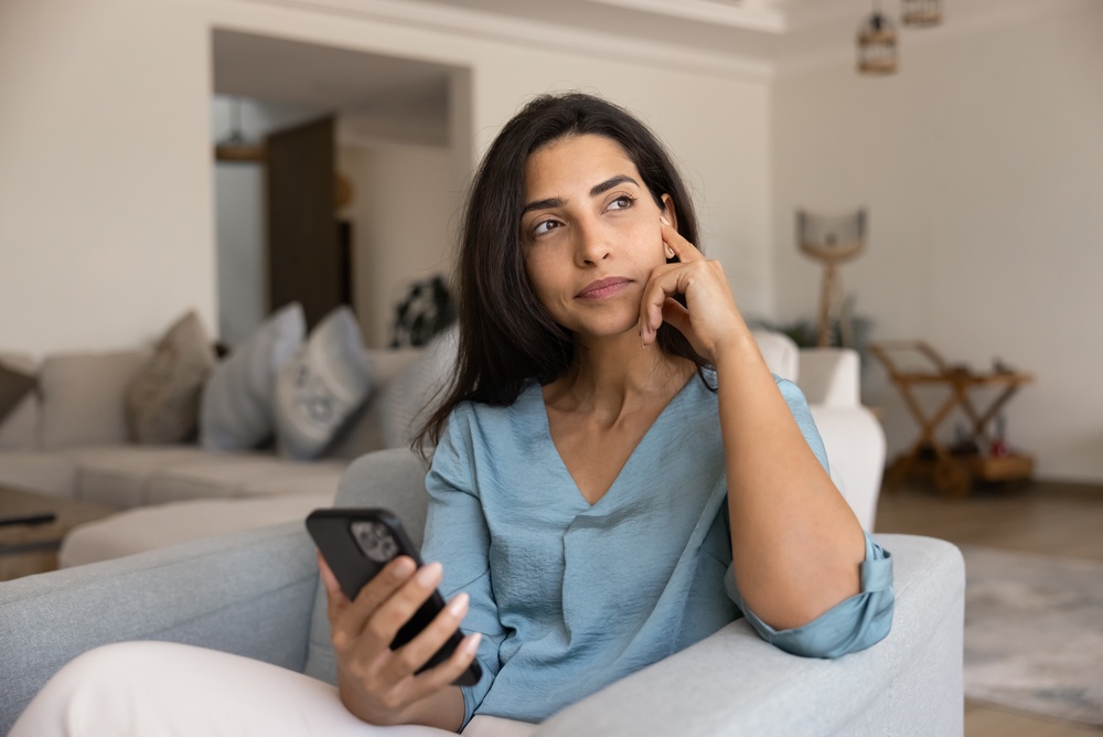 Woman sitting in a gray armchair at home holding her phone, resting her head on her hand and looking off to the side.