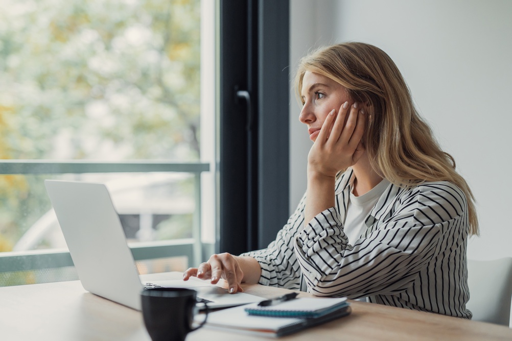 Woman at a laptop by a window with autumn trees outside, resting her cheek on her hand while she thinks.